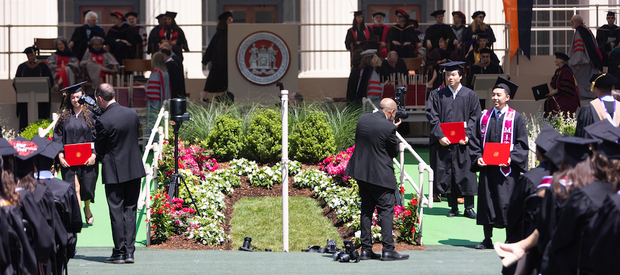 Image of graduates walking down the ramp from the stage with their diplomas in hands. They are being photographed by photographers dressed in black.