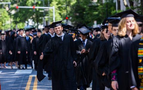 A graduate in regalia gives a thumbs-up from the procession line.