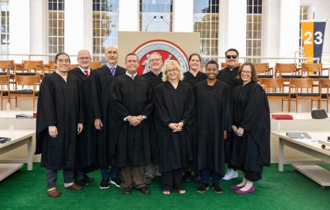 Image of the registrar's office smiling in front of the podium on the stage. They are all wear black regalia robes and smiling at the camera.