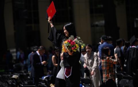 Image of a graduate after the Undergraduate Ceremony smiling and holding up her diploma and holding a bouquet of flowers