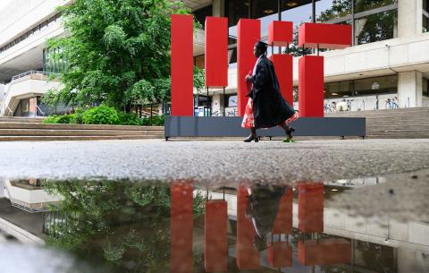 Image of a graduate in their regalia walking by a large sculpture that reads MIT